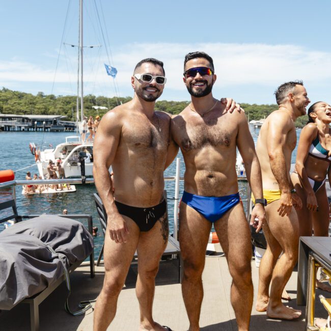 Two men smiling on a boat; one in black swimwear and sunglasses, the other in blue swimwear and sunglasses. More people are in the background, some on the boat and some in the water. It's a sunny day with clear skies.