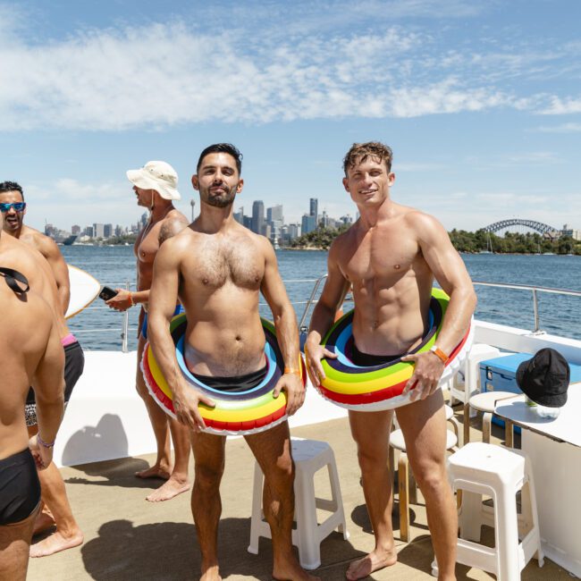 Two men are posing on a boat deck, each wearing a colorful inflatable ring around their waist. They're shirtless, wearing swim trunks, and smiling. The background shows a city skyline and a harbor under a clear blue sky.