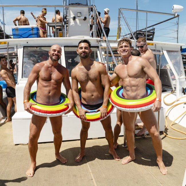 Three men in swimwear stand on a boat deck, each wearing a rainbow-colored inflatable around their waists. Other people are visible in the background, along with boats and flags. It's a sunny day.