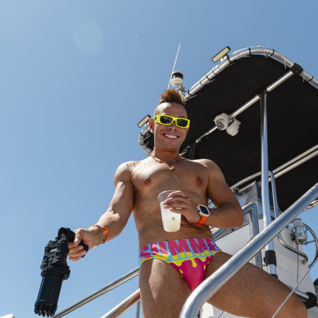 A person in colorful swimwear and sunglasses stands on a boat's steps under a clear blue sky. They are holding a drink in one hand and a black object in the other, smiling brightly. The boat has metal railings and a canopy.