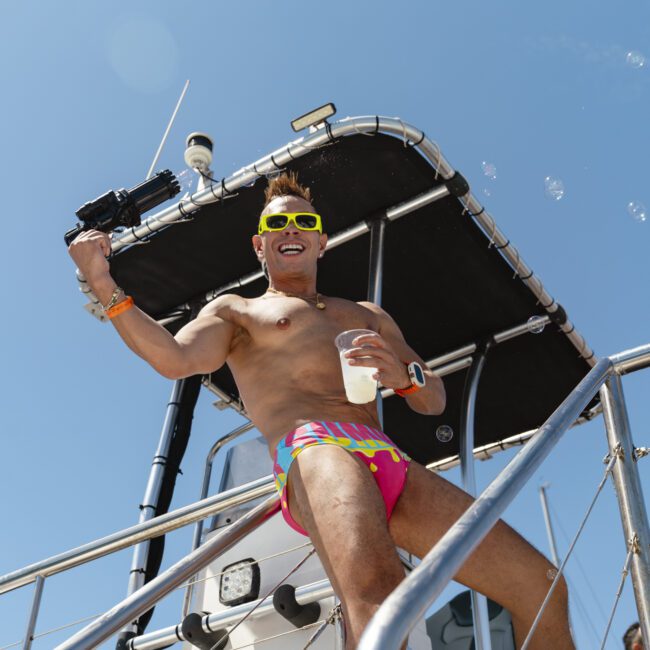 A man in colorful swim trunks and neon sunglasses stands on a boat, smiling and holding a water gun and a cup. The sky is clear and blue, and bubbles float around him. Other people are partially visible on the boat.