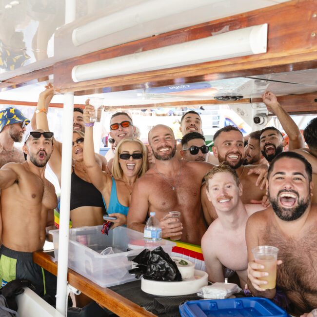 A group of smiling people gathered on a boat, many wearing swimsuits and sunglasses. They are raising drinks and posing for the camera, creating a lively and cheerful atmosphere.