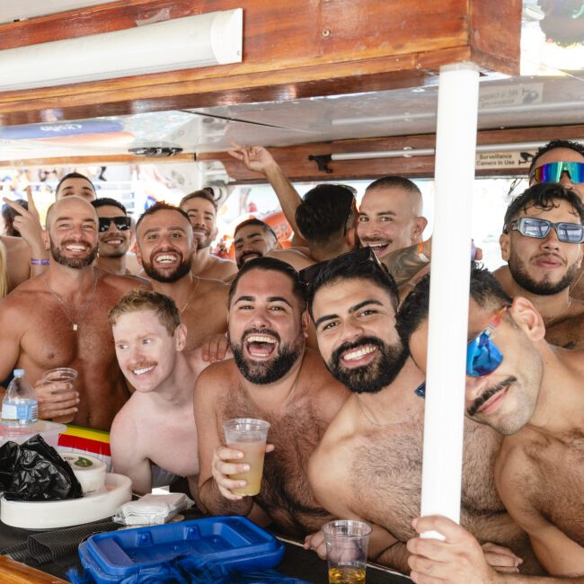 A group of people smiling and holding drinks on a boat. They are gathered closely together, some wearing sunglasses and swimwear, appearing to enjoy a sunny day on the water. Some have various accessories like hats and necklaces.