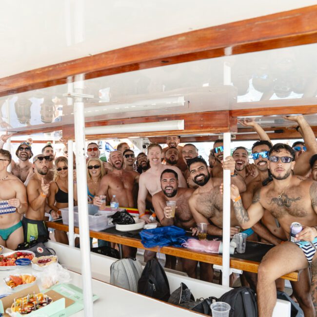 A group of people in swimsuits are gathered closely on a boat, smiling and raising their arms in celebration. The boat table is covered with food and drinks. The background shows water, indicating they are likely on an outing or party at sea.