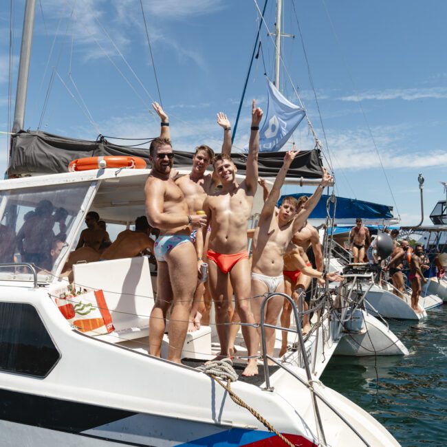 Four men in swimwear are standing on the deck of a boat, raising their hands and smiling. The boat is docked among other boats on a sunny day, with a clear blue sky overhead.