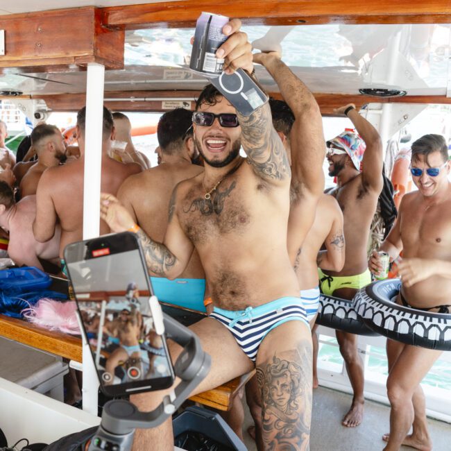 A group of people in swimwear enjoying a lively party on a boat. A smiling man with sunglasses and tattoos raises a drink. Someone records the scene on a smartphone, capturing the festive atmosphere. Others hold drinks and wear inflatable pool toys.