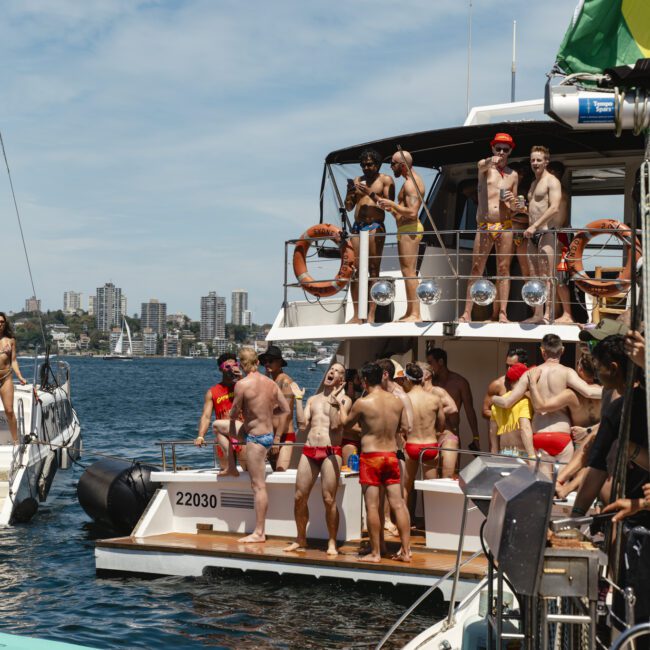 People are enjoying a sunny day on a boat wearing swimwear. Some are standing on the upper deck, while others are gathered on the lower deck. The boat is surrounded by water, and a city skyline is visible in the background.