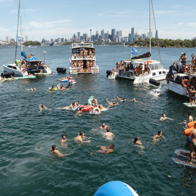 A group of people swim and relax in a harbor, surrounded by several boats. The skyline of a city and a bridge are visible in the background. The atmosphere is lively and festive under a clear blue sky.