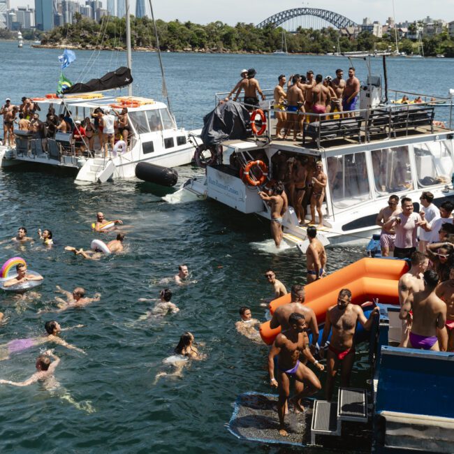 People are enjoying a boat party on the water. Some are swimming, others are on boats with inflatables. The scene is lively with many people relaxing and socializing. In the background, a city skyline and a bridge are visible.