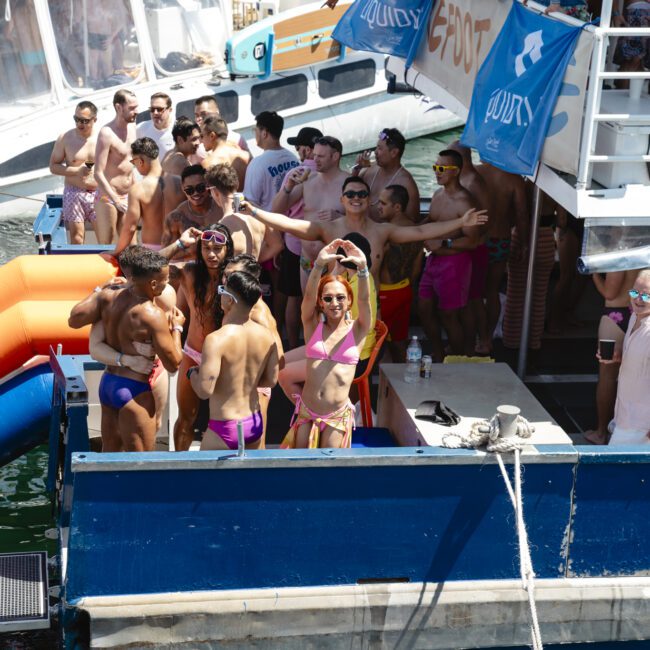 A lively crowd of people in swimsuits enjoying a party on a boat. Some are dancing, others are in conversation, and one person in the foreground is forming a heart shape with their hands. Inflatable floats are visible by the water.