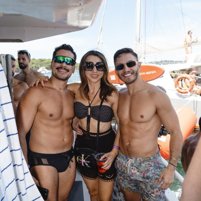 Three people posing on a boat deck under a clear sky. The two men, shirtless, flank a woman in a black swimsuit. They are smiling, with a lifebuoy and part of a sailboat visible in the background.