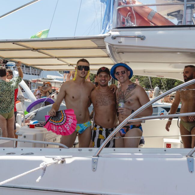 Three people in swimwear and sunglasses pose cheerfully on a boat. One holds a vibrant pink fan. Other individuals are visible in the background, enjoying the sunny day on the water.