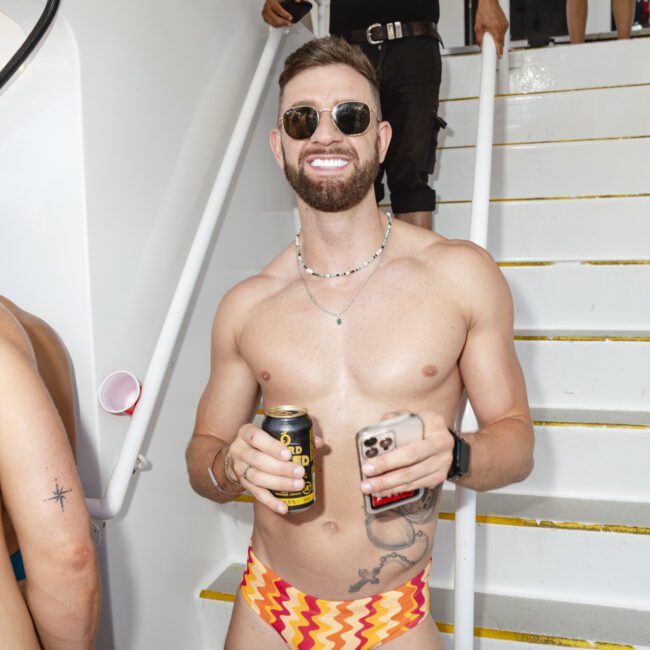 A shirtless man in sunglasses and colorful swim briefs holds a can and a smartphone while smiling on a boat. He stands in front of stairs as people walk behind him.