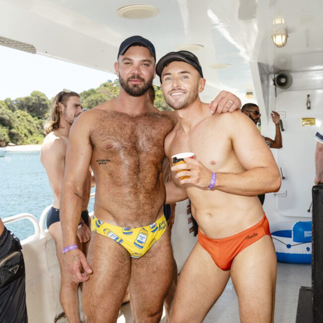 Two men in swimwear smiling on a boat. The man on the left wears a yellow swim brief and a cap, while the man on the right wears an orange swim brief and holds a drink. Others are visible in the background, with a view of the water and trees outside.