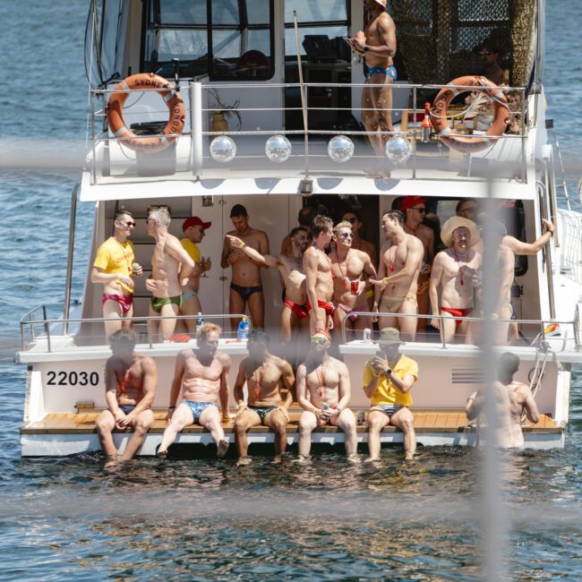 A group of people in swimwear are gathered on the deck of a boat, some sitting and some standing. The boat is on a body of water, with greenery visible in the distance. A few people are also in the water near the boat.