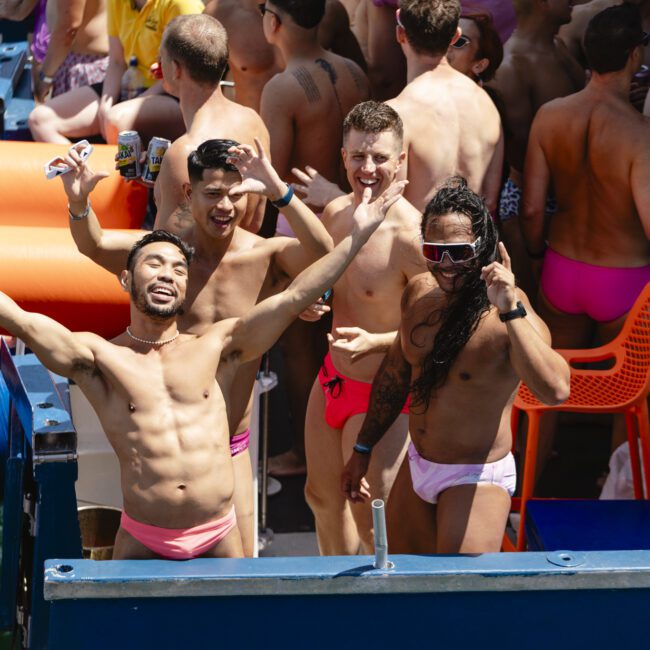 A group of people in swim trunks are enjoying a sunny outdoor party on a boat. Some are standing, waving, and smiling at the camera, while others are relaxing and socializing. Bright colors and festive atmosphere are evident.