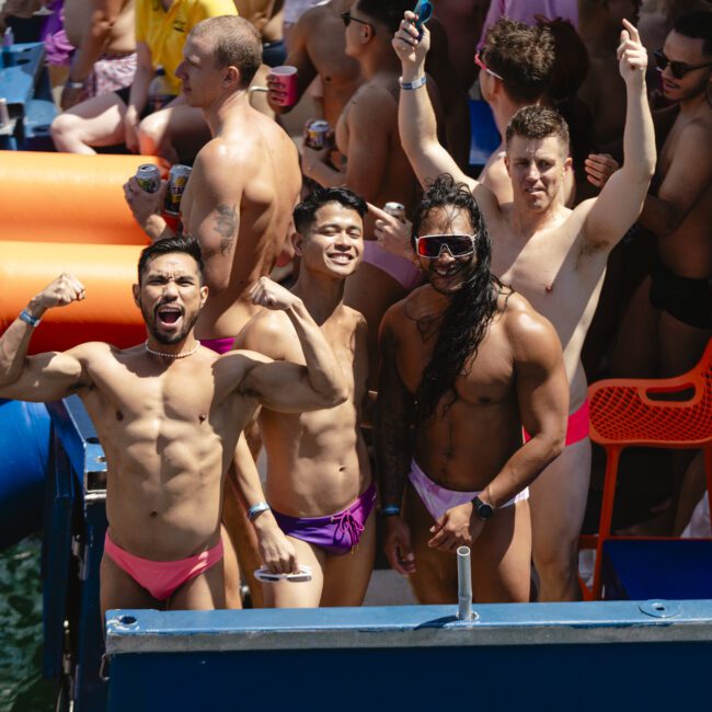 A group of four men in swimwear enjoy a lively outdoor party on a boat. They are smiling and posing energetically, with some flexing their muscles. Other partygoers and inflatable items are visible in the background near the water.