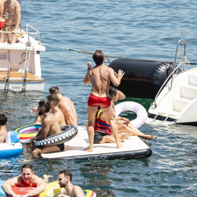 A group of people enjoying a sunny day on the water, using inflatable floaties and a raft. Some are standing or sitting, while others are lounging on a nearby boat. The scene is lively and colorful.