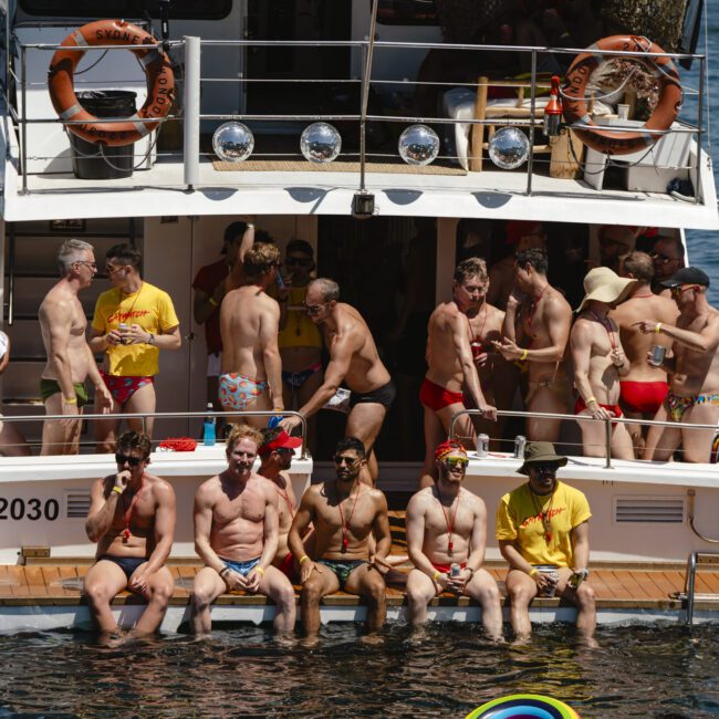 A group of people in colorful swimwear are gathered on and around the back of a boat on a sunny day. Some are sitting on the edge with their feet in the water, while others stand chatting on the deck. The water is bright blue.