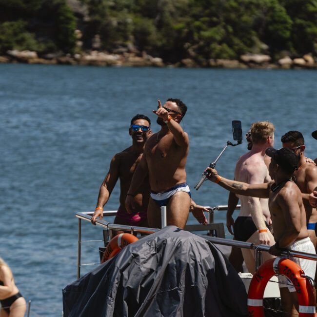 A group of people enjoying a sunny day on a boat, with one man taking a selfie using a phone on a stick. They are surrounded by water, with trees and rocks in the background. Most individuals are wearing swimwear and sunglasses.