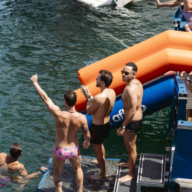 Three men stand on a platform by the water, wearing swim trunks. One man raises his hand in a thumbs-up gesture. There is an inflatable slide nearby, and other people are visible in the water and on boats.