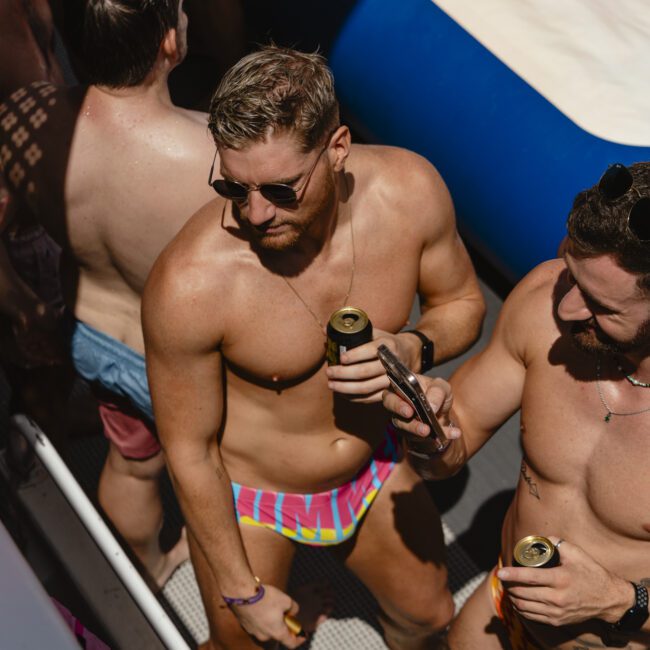 People in vibrant swimwear gather on a boat under the sun. Two individuals in sunglasses hold drinks and converse. The atmosphere is relaxed and lively, with shadows and sunlight creating patterns on their skin.