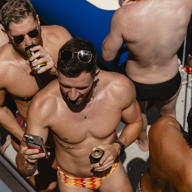 A group of men in swimwear enjoying a sunny day on a boat. One man in patterned swim trunks holds a phone, while another nearby sips a drink. Inflatable pool floats are visible in the background.