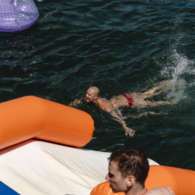 A person swims in a body of water, wearing red swim trunks, near an inflatable seahorse. Another person stands on an orange and white inflatable slide in the foreground. The water is dark and wavy.