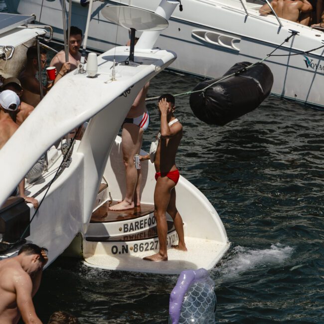 People in swimsuits enjoy the sun on boats and rafts on the water. A group stands on the deck of a white boat, while others are on a floating inflatable in the shape of animals. The scene is lively with clear skies and calm water.