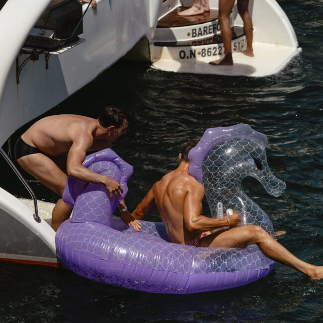 Two people are on a large purple inflatable seahorse in the water near a boat. One person helps the other climb onto the float. Several others are on the boat, some wearing swimsuits. The scene is lively and set in sunny weather.