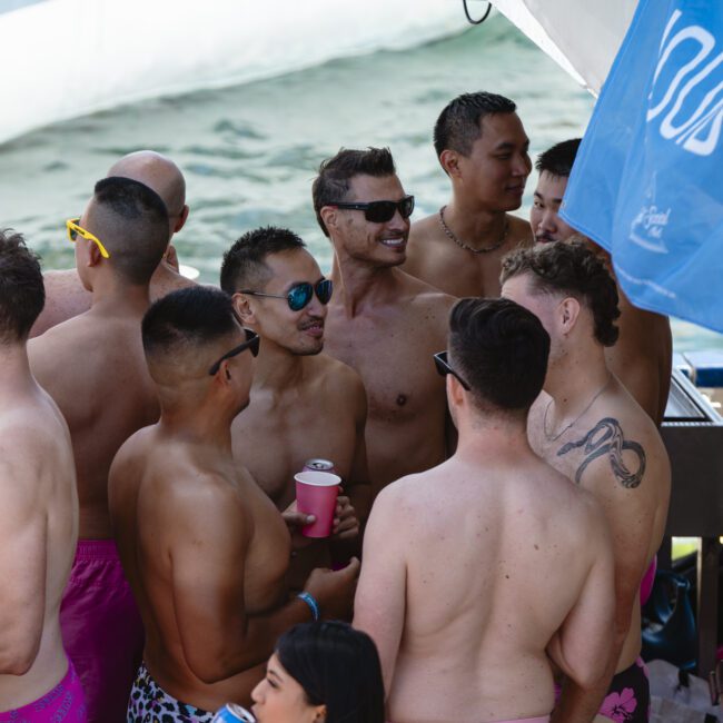 A group of shirtless men, many wearing sunglasses and swim trunks, are gathered on a boat under a blue canopy. They appear to be socializing and enjoying a sunny day. One person is holding a pink drink can.