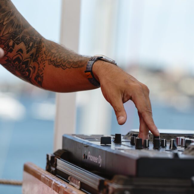 A person with a tattooed arm adjusts knobs on a DJ controller with a blurred outdoor background.