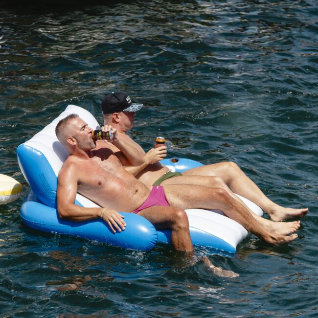 Two men float on a blue and white inflatable lounge in the water, both drinking from bottles. One wears a black cap and black trunks, while the other wears purple trunks. They are surrounded by gently rippling water.