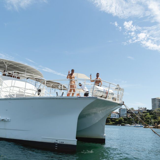 Two people waving from the deck of a large white yacht in a sunny harbor. The water is a clear blue, and the sky is dotted with clouds. Buildings and greenery are visible on the shore in the background.