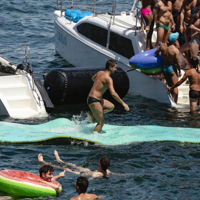 People enjoying a sunny day at sea. A man in black swimwear runs on a floating mat between boats. Others swim nearby, with one person holding a watermelon-shaped float. More people gather on the boats, all wearing swimwear.