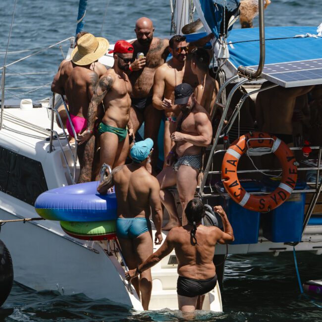 A group of men in swimwear gather on a sailboat and in the water. Some are standing on the deck, while others are in the water near the boat. Inflatable rings and a lifebuoy are visible, with the background being a sunny day at sea.