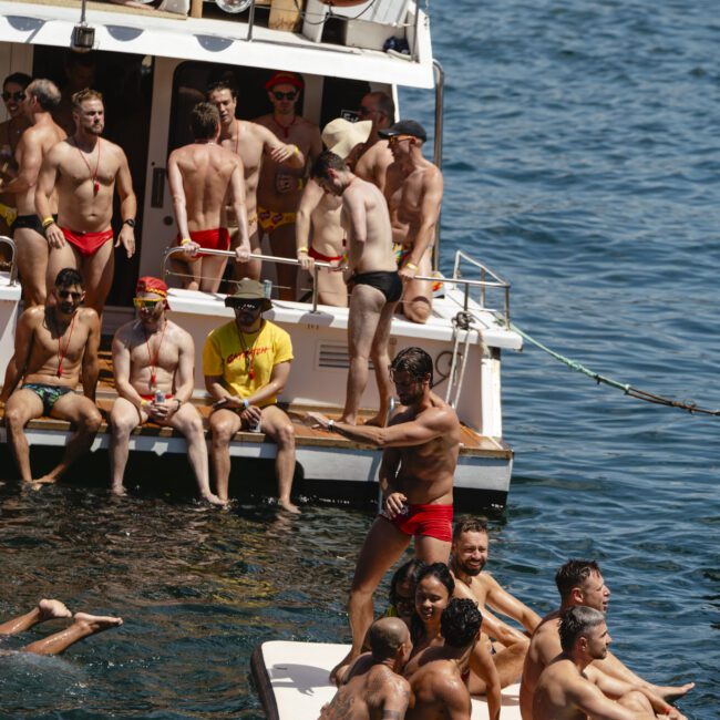 A group of people in swimwear enjoy a sunny day on a boat and in the water. Some are lounging on a float, while others swim or stand on the boat deck. The scene is lively and relaxed, surrounded by clear blue water.