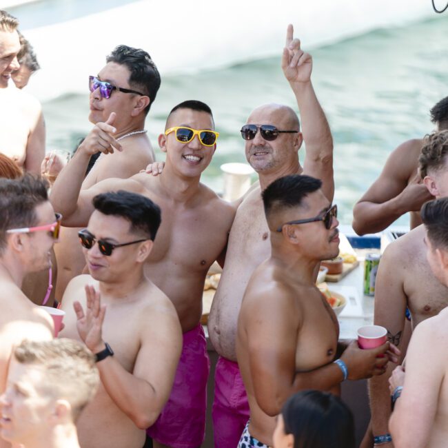 A group of shirtless men in sunglasses and shorts are gathered on a boat, enjoying a party. Some hold drinks and smile, while one man raises his arm in excitement. The backdrop shows water and part of a boat.