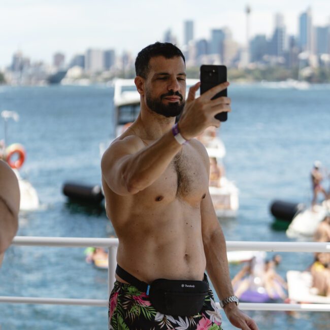 A shirtless man with a beard takes a selfie on a boat. He wears floral swim shorts and a black waist bag. The background shows a body of water with floating platforms and a city skyline in the distance.