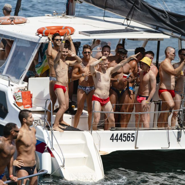 A group of men in swimwear, mostly speedos, are on the deck of a boat on the water. Some are standing, waving, and smiling. Other boats with people are nearby, and the weather seems sunny and clear.