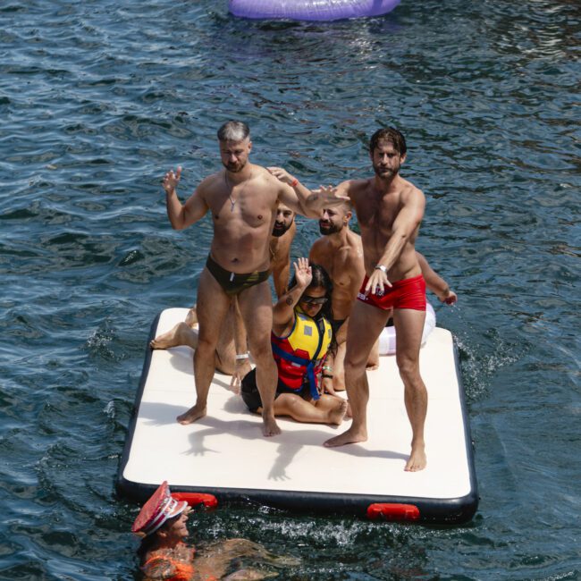 A group of men in swimwear stand and sit on a floating platform in the water, posing and enjoying the sun. In the background, a person floats in a purple inflatable unicorn near a boat, and another person swims nearby wearing a colorful hat.