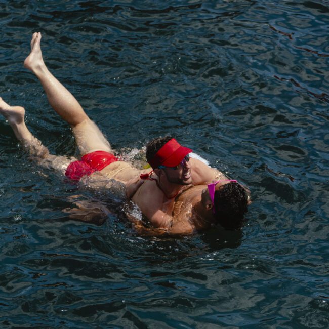 Two people in a lake, one carrying the other on their back. Both are wearing red swimming trunks, with one in a red visor and the other in a pink headband. They appear to be laughing and enjoying the water.