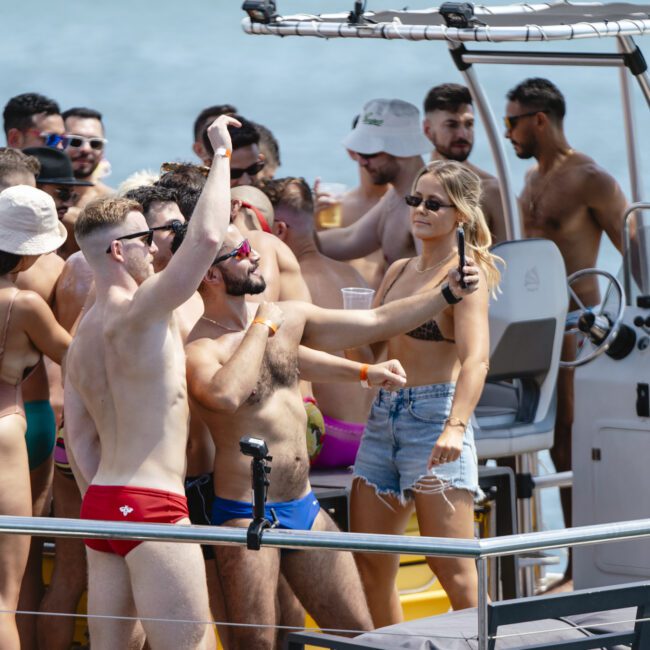 A lively group of people in swimwear are taking selfies and enjoying drinks on a boat. The scene is festive, with men and women interacting under sunny weather, with the boat in motion on the water.