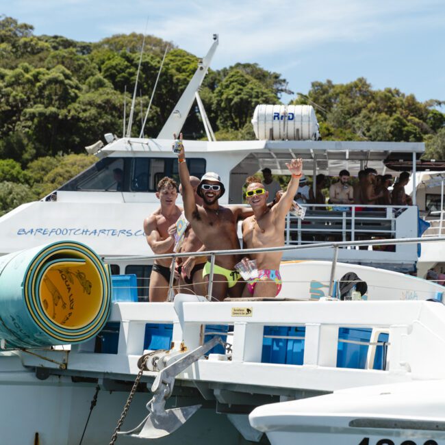 Three people in swimwear stand on a boat, smiling and waving at the camera. Another boat and lush greenery are in the background. The sky is clear and sunny.