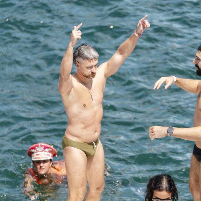 A man in a speedo stands on a platform above the water with arms raised, next to a woman in a life vest and another man. A person with a hat floats in the water nearby. The background shows rippling water.