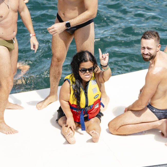 A group of people in swimwear are on a floating platform. A woman wearing a yellow life vest sits in the center, smiling and gesturing with her hand. The background shows water, suggesting a recreational setting.