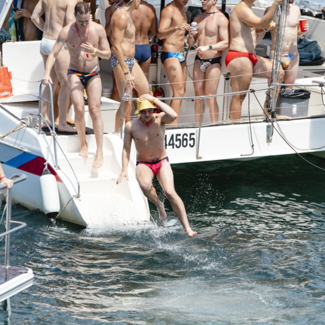 A group of people in colorful swimwear are on a boat. Some are standing and talking, while one person in a red swimsuit and yellow hat is jumping into the water. Background shows the sea on a sunny day.