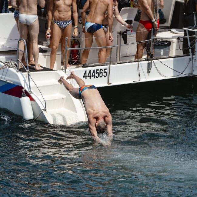 A group of people in swimsuits are on a boat. One person is diving into the water while others stand on the deck, some holding drinks. The water is calm, and it's a sunny day. The boat has steps leading into the water.