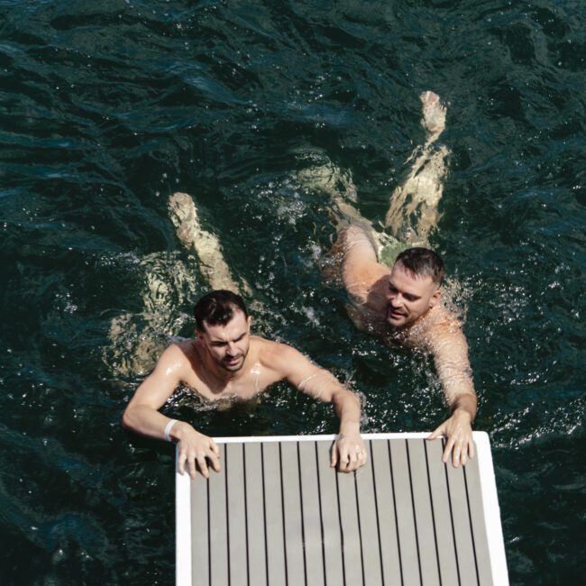 Two men swimming in dark green water, holding onto the edge of a floating platform. Both are shirtless, with short hair, appearing relaxed and enjoying the water on a sunny day.