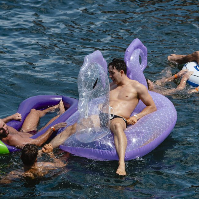 A man lounges on a transparent inflatable sea serpent float in the water, surrounded by friends on other floats. They appear to be enjoying a sunny day at the lake.